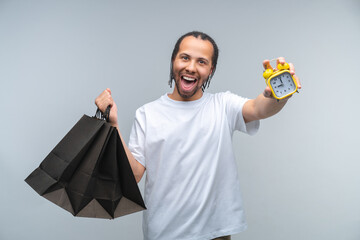 Studio portrait of euphoric young African American man customer holding a bunch of shopping bags and reaching and alarm clock in his hand to the camera
