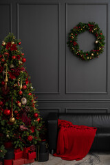 Cozy and elegant Christmas scene set against a dark gray paneled wall. A beautifully decorated Christmas tree adorned with red and gold ornaments and baubles. A black leather sofa is partial visible