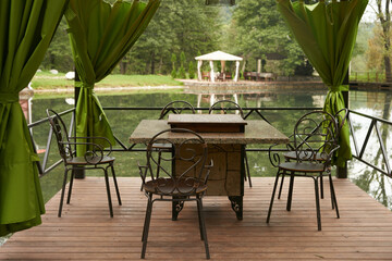 Summer cafe on the shore of a picturesque blue lake. Table and chairs on a wooden deck - a pergola with textile curtains. Trees beautifully reflected in the water.