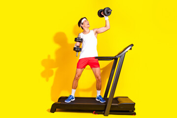 Young man in activewear joyfully lifting dumbbells on treadmill against vibrant yellow backdrop