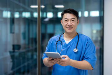 Asian man doctor wearing blue scrubs smiles while holding tablet in modern medical facility....
