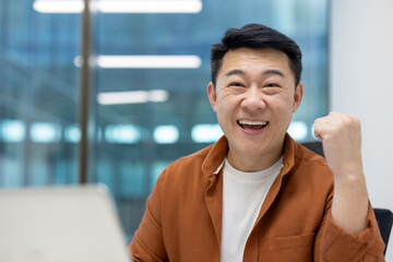 Asian man businessman expresses joy and success in office. His smile reflects motivation and confidence, emphasizing professional achievements and positive attitude. Success celebration is evident.