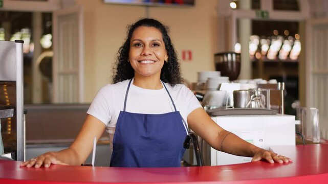 Confident Latina shop owner smiling behind store counter in apron. Proud small businesswoman posing at retail counter