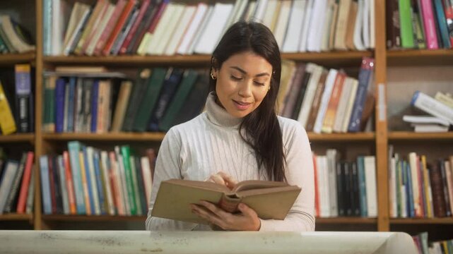 Smiling woman reading an old book while standing in front of shelves in a cozy library. Calm reader enjoying vintage literature in a peaceful bookstore setting.