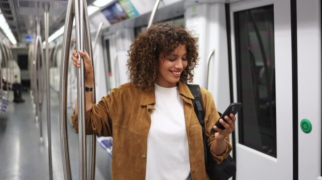Young woman stands on a subway train, holding a smartphone and browsing social media with a smile.  Woman browsing social media on subway in slow motion