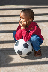 Sad African child, a soccer ball nestled between his legs, fists covering his mouth,
