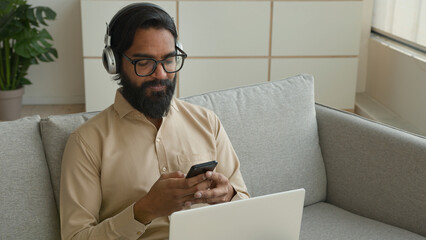 Arabian Indian businessman man multitasking working with devices at home muslim guy in headphones talk video chat meeting call on laptop typing cell phone message use mobile app online communication