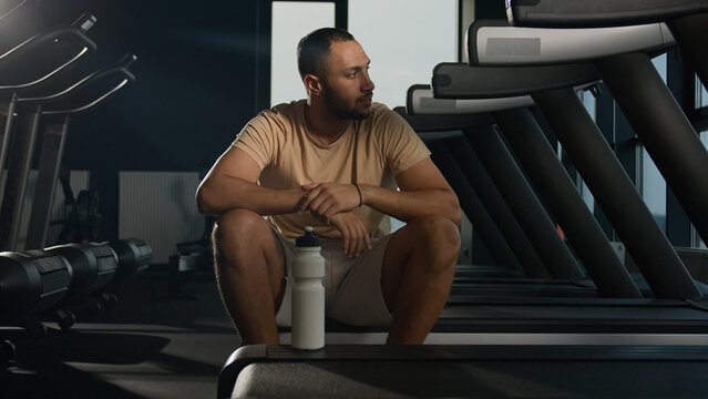 Tired African American man sit on treadmill relax after hard physical workout cardio training in fitness club gym exhausted sweaty sportsman runner thirsty drink water sport bottle healthy lifestyle