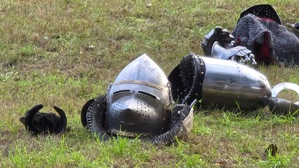 Medieval armor lying on grassy ground, including a helmet with a visor, chainmail, and metal gauntlets. The scene suggests a pause or aftermath of a historical reenactment or battle display.