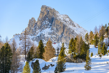 Majestic snow-capped rocky peak overlooking a ski resort in the Alps on a clear winter day. A red aerial tramway going up is visible against the mountain face.