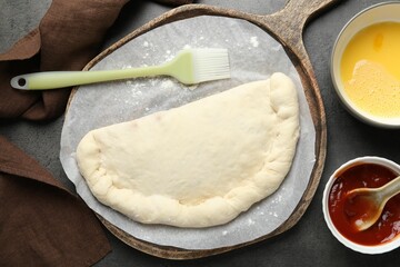 Uncooked calzone pizza, sauce and egg yolk on grey table, flat lay