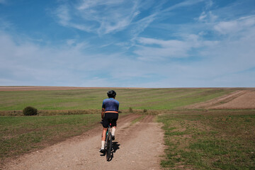 Obraz premium Cyclist rides gravel road across vast, idyllic countryside under a vibrant blue sky. Perfect for themes of adventure, freedom, and rural exploration.