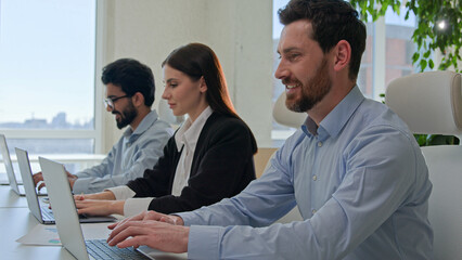 Row of diverse smiling business people businessmen and businesswoman typing on laptops professional colleagues team of technical support staff members work on desktop computers co-working office space