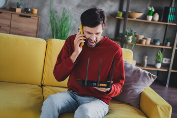 Young man in casual pullover frustrated with home internet issues, holding router and phone indoors...