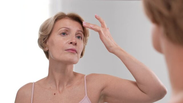 Beautiful woman in her 40s critically examines her appearance in the mirror, upset and anxious about skin imperfections and wrinkles. Slo mo