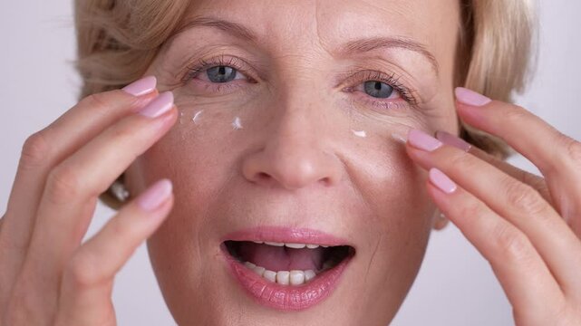 A gorgeous woman in her 50s applying eye cream as she smiles looking in the mirror. Closeup portrait. Aging and thriving wellness