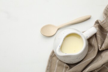 Condensed milk in jug and spoon on white marble table, top view. Space for text