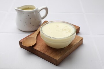 Tasty condensed milk in bowl and jug with spoon on white tiled table, closeup