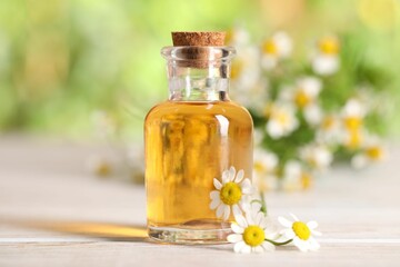 Bottle of essential oil and chamomile flowers on white wooden table against blurred background, closeup