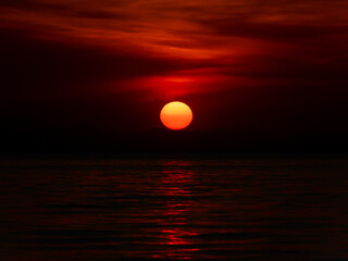 A stunning sunset paints the sky in deep reds as the sun sinks below the horizon, reflecting on calm waters and creating a serene atmosphere in the evening.