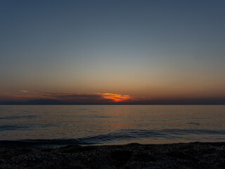 A breathtaking sunset casts warm hues across the sky while the calm ocean reflects golden and orange tones. The shoreline remains tranquil, evoking serenity during dusk.