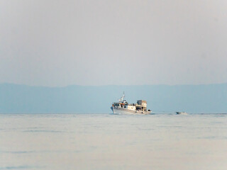 A fishing boat moves slowly through calm waters, with a hazy skyline in the background. The atmosphere is peaceful, highlighting the solitude of the early morning.
