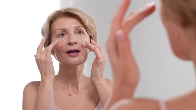 A positive woman in her 50s using moisturizing cream as she touches her face while finger-tipping. Self love and skincare rituals.