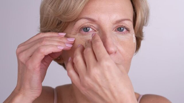 A middleaged female looking into the mirror, applying eye patches and smiling. Gracefully aging female takes care of her skin and beauty