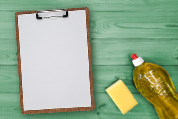 Clipboard checklist Blank, bottle of detergent, kitchen sponge on green table. Sheet of paper on Clip board with Place for text, messages, list, flat lay. Planning housekeeping spring cleaning.