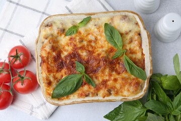 Delicious cooked lasagna in baking dish, basil and tomatoes on light grey table, flat lay