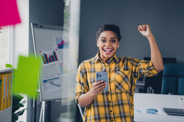 Photo of attractive young woman winning hold device wear yellow plaid shirt comfortable modern office loft room interior indoors workspace