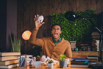 Young man frustrated with homework, sitting in room full of books, expressing stress and irritation while studying at home in the evening.