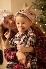 A beautiful mother and child in a checkered shirt are having fun near the Christmas tree. A woman and a child in checkered pajamas are satisfied and happy on a Christmas background