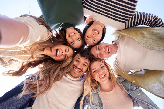 Selfie portrait from below of diverse generation z group friends posing smiling in circle looking at camera outdoor. Happy multiracial young people having fun embracing together sunny day. Low angle