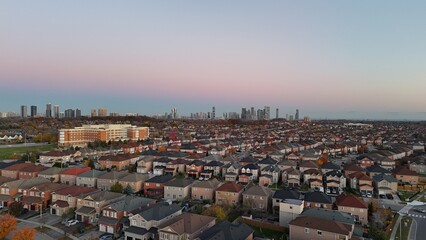 Obraz premium A scenic aerial view of a suburban neighborhood during sunset, with rows of colorful houses leading to a distant city skyline under a gradient sky transitioning from pink to blue. A vibrant blend of u