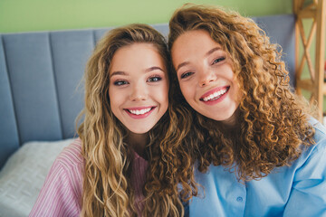 Charming two girls with beautiful smiles relaxing at home in light bedroom interior