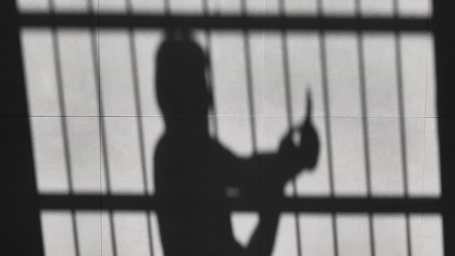 shadow of prisoner man using phone behind the jail bars reflected on the floor,silhouette of kidnapped person using mobile device to communicate outside