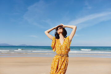 Woman in yellow dress posing on sandy beach with hands on head in contemplation and relaxation