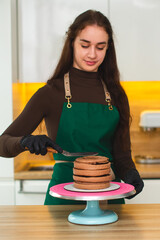 Close-up. A young confectioner girl in a green apron covers a cake with chocolate cream using a culinary spatula.