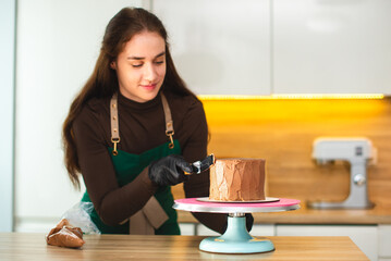 The smiling female pastry chef smooths the surface of the chocolate cake with a culinary spatula. White kitchen on background.