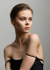 Elegant young woman in a black dress with a thoughtful expression, showcasing natural beauty against a soft gray background. Fashion and portrait photography concept.