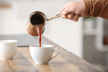 Female hand pouring hot brewed coffee from cezve in cup on table in kitchen