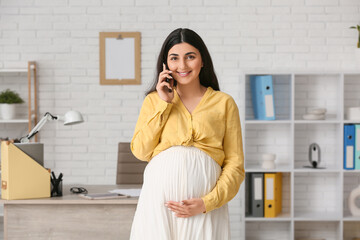 Beautiful pregnant businesswoman talking with mobile phone near workplace in office