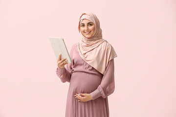 Beautiful pregnant Arabian businesswoman in hijab using tablet on pink background