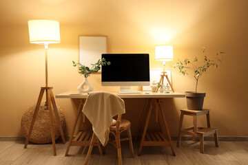 Stylish workplace with modern computer and glowing lamps near yellow wall in office at night