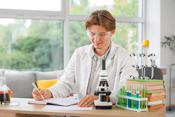 Male student in lab coat with notebook and microscope studying at home