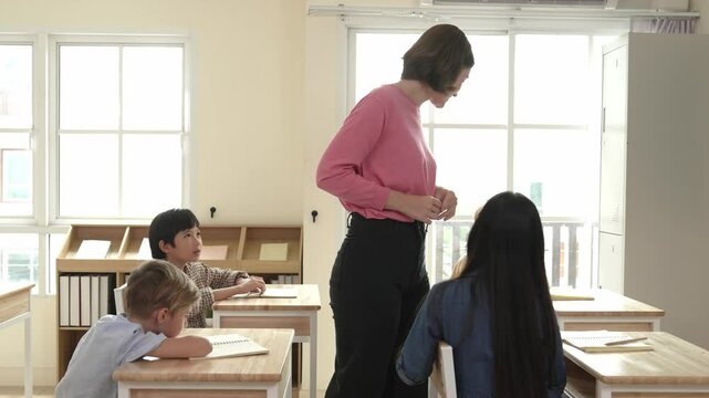 Caucasian teacher walking and check student homework at classroom while diverse children doing classwork at classroom. Diverse smart student lecturing and taking a note at elementary school. Pedagogy.