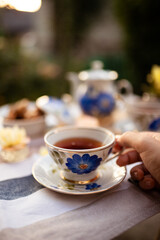 Woman's hand holds a cup of tea. A table is served and decorated with a beautiful porcelain set of dishes decorated with blue flowers.
