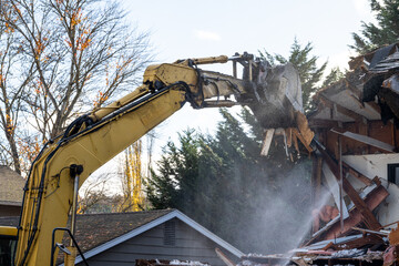Large yellow excavator starting demolition on a house, workman spraying water to keep dust down,...