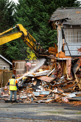 Large yellow excavator starting demolition on a house, workman spraying water to keep dust down,...